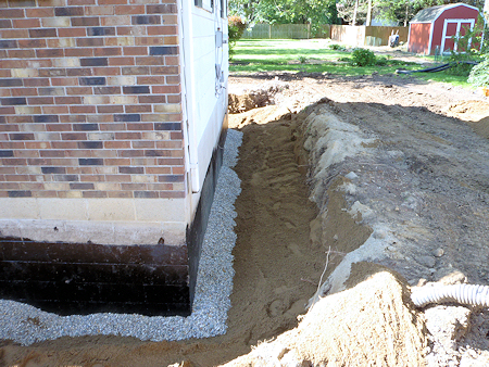 Excavated soil and drainage aggregate beside a brick foundation during exterior water-control work